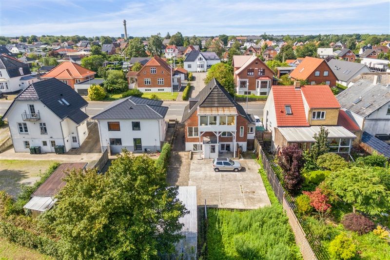 Have med grønne træer og buske. To biler parkeret foran husene. Huset i centrum med garage og en bil parkeret. Træer og
