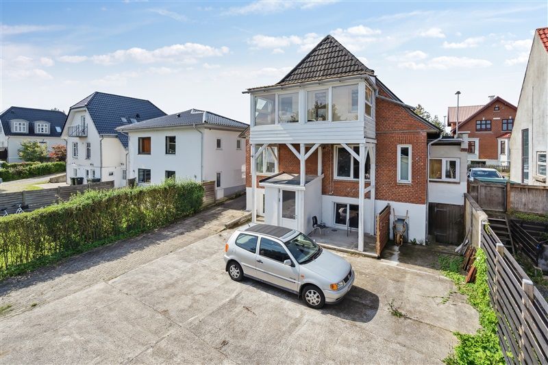 Afkast 6,05% Haderslevvej 127, 6000 Kolding En sølvfarvet bil parkeret foran et hus med terrasse, omgivet af grønne hegn og