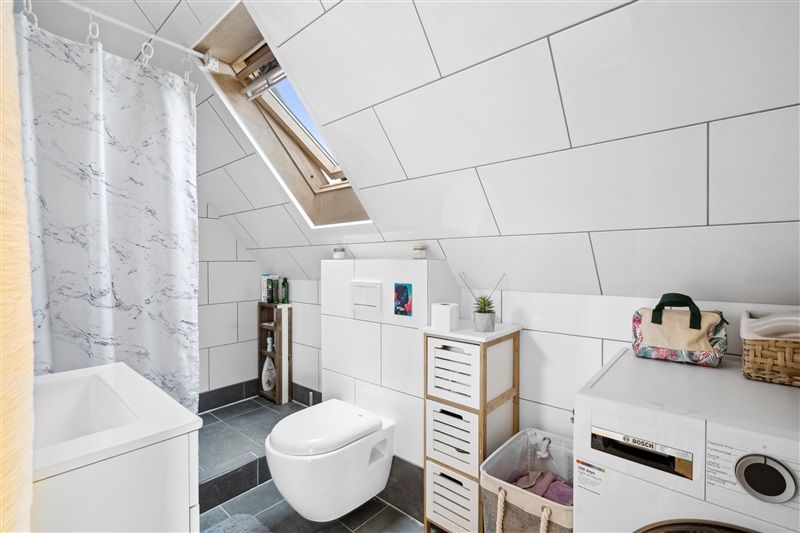 Toilet med badekar, brusegardin og højt vindue. Hvid gulv, toilet, vask og toiletpapir. To træhylder med