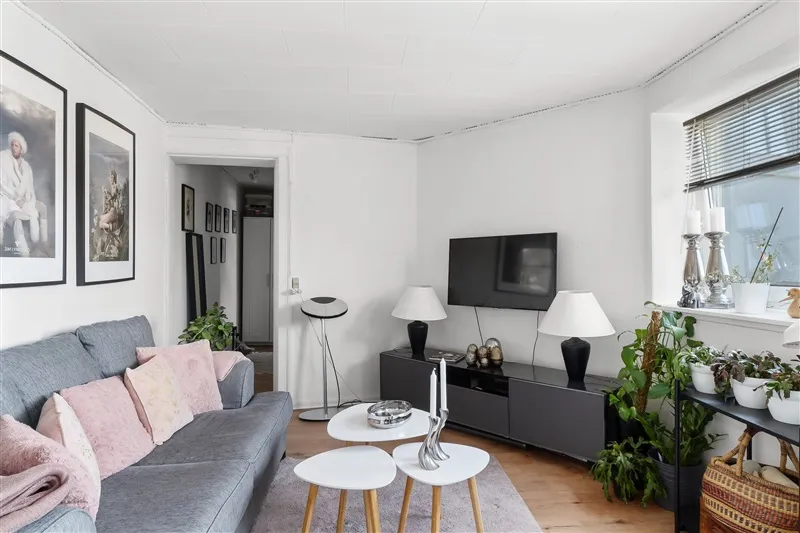 Lyst og ryddeligt hus med en grå sofa, en tv-enhed, og to små stolpeborde med hvide lamper og et tv på væggen