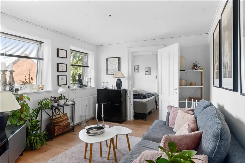 Lyst og rummeligt værelse med dobbeltseng og hyggeligt møbelsæt. Elegant indretning med plante og lyse farver.