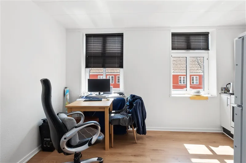 Minimalistisk arbejdsværelse med høj højde og hvide vægge, computer på bordet, skrivebordsstol og køkken med lyse skuffer og