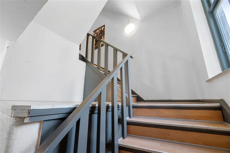 En tre-trins trehøj hvid trappe med skrå gulv og metalgesims. Sort blyant hænger på væggen og en lysplet tændes.