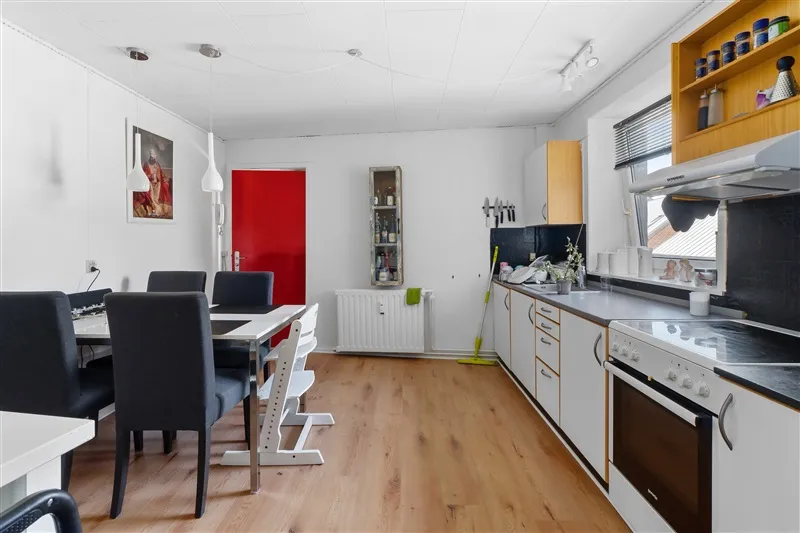 Køkken med højloft, lyse lamper, lyserødt dørblad og trægulv med en hvid spisestue.