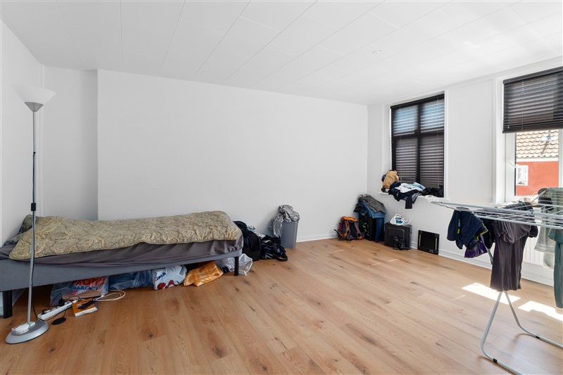 Et tomt værelse med en enkel seng og en stående lampe samt en klædeskabet stander med klæder hængende på den