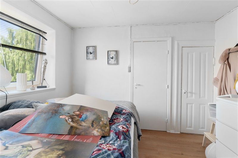 Soveværelse med hvide vægge og trægulv, lysegardiner og udsigt til træer, og et stort billede med kunst på sengen