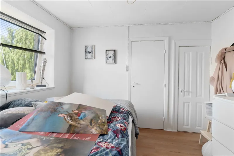 Soveværelse med hvide vægge og trægulv, lysegardiner og udsigt til træer, og et stort billede med kunst på sengen