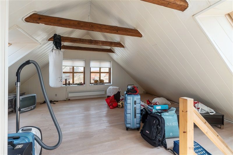 Loftsrum med to vinduer og træloft. To kuffert, to poser og en støvsuger på gulvet. Et tv og en radiator på loftsgulvet