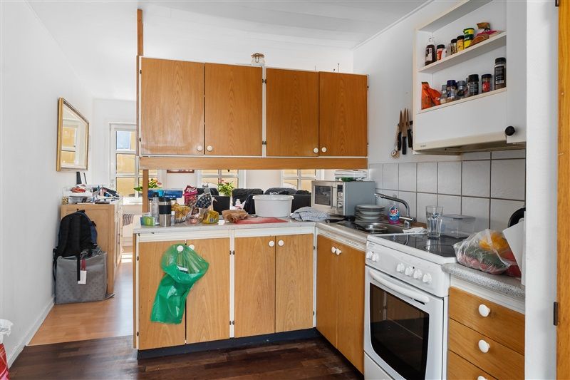 Køkken med høje træskabe, stående komfur, indbyggede ovne, plastposer og glas på loftshylder, gryder og glas på en bordplade