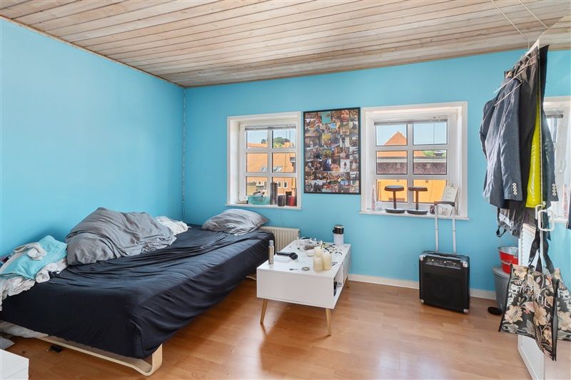 Et enkelt soveværelse med højloft og lyseblå vægge. På gulvet står en seng med svedige og et hvidt sengehoved. Et hvidt bord