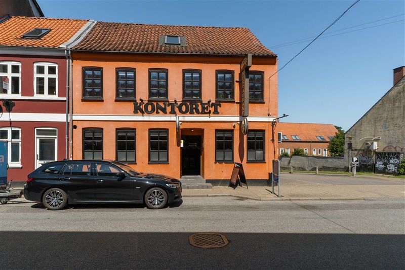 En sort BMW står parkeret foran en orange bygning med teksten Kontoret på facaden. Bygningen har mange vinduer og et rødt