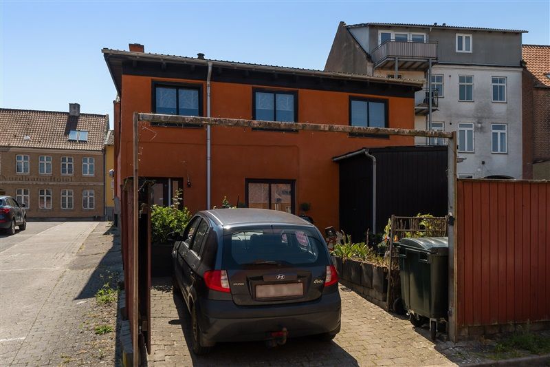 En lille, grå bil parkeret foran en orange og hvid bygning med en port og en have på siden