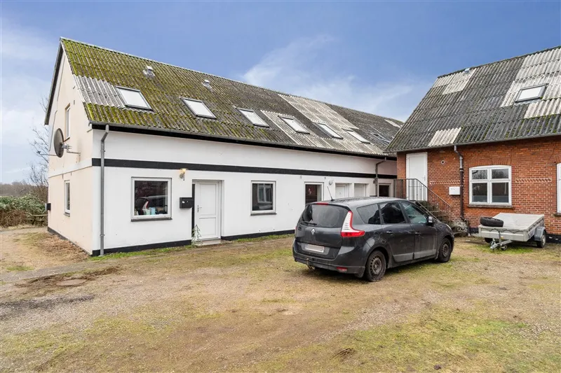 En sølvfarvet Renault parkeret foran en hvid og brun bygning med røde mursten og en trailer med et dæk bagved.