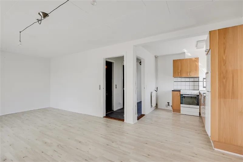 En tom lejlighed med hvide vægge og lyse trægulve, lysekroner hængende fra loftet og et køkken med indbygget ovn og høje