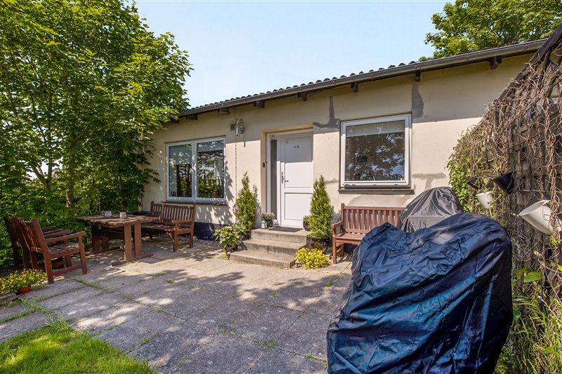 Have med bord, bænke, græsplæne og beplantning omkring hus med tre vinduer og hvid dør