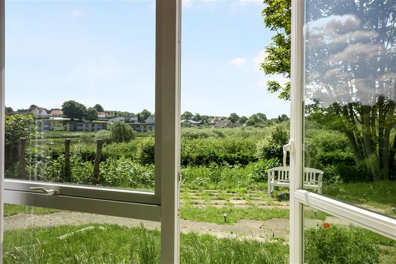 Usynlig udsigt fra et vindue mod en gård med græs og træer, hvor en hvid bænk står udendørs