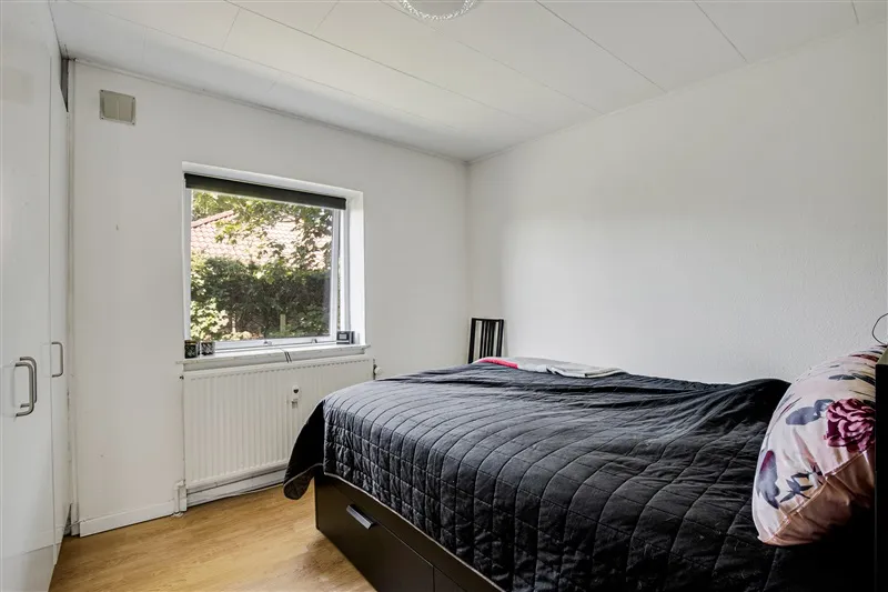 Soveværelse med hvidt loft, hvide vægge og trægulv, sort sengelinned og pude med blomsterprint og radiator med hvide væggen