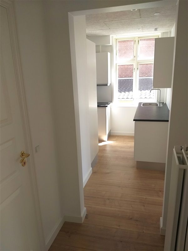 Moderne køkken med en stor vindue, lyse trægulv og en hvid overdel. Vinduet giver udsigt til en gårdhave.