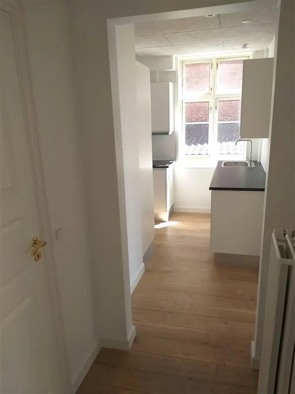 Moderne køkken med en stor vindue, lyse trægulv og en hvid overdel. Vinduet giver udsigt til en gårdhave.