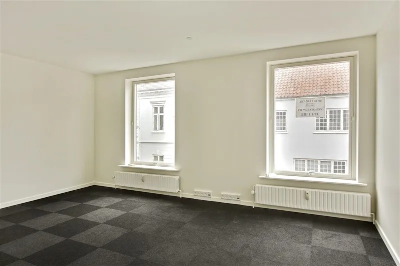 To vinduer med hvide rammer og tætsiddende persienner i hvidt og gråt rum med hvide vægge og gulv. Radiatorer under vinduer