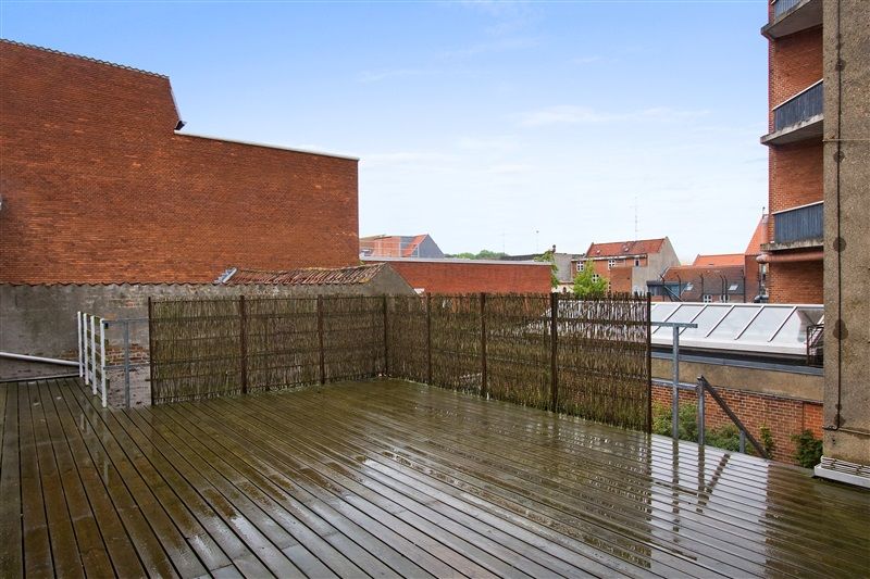 Terrasse med trægulv og lysgrå metalhegn omkring. Bagved er en høj bygning med røde mursten og en hvid tagdækning. På