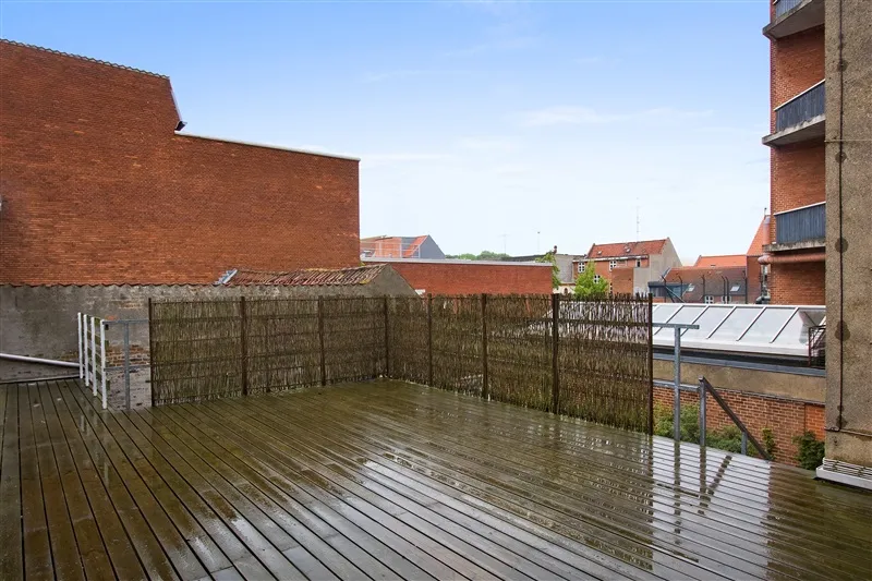 Terrasse med trægulv og lysgrå metalhegn omkring. Bagved er en høj bygning med røde mursten og en hvid tagdækning. På