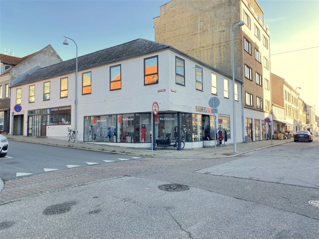 To høje bygninger med store vinduer, hvor det står Red Barnet. En cykel står parkeret udenfor. Parkerede biler og gågade i
