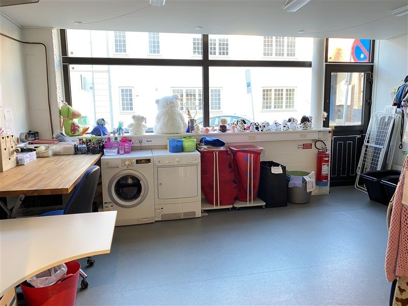 Et værelse med vasketøjsmaskine, tørretumbler og diverse kuverter og søjler med tøj. Et bord med diverse ting og et skab med