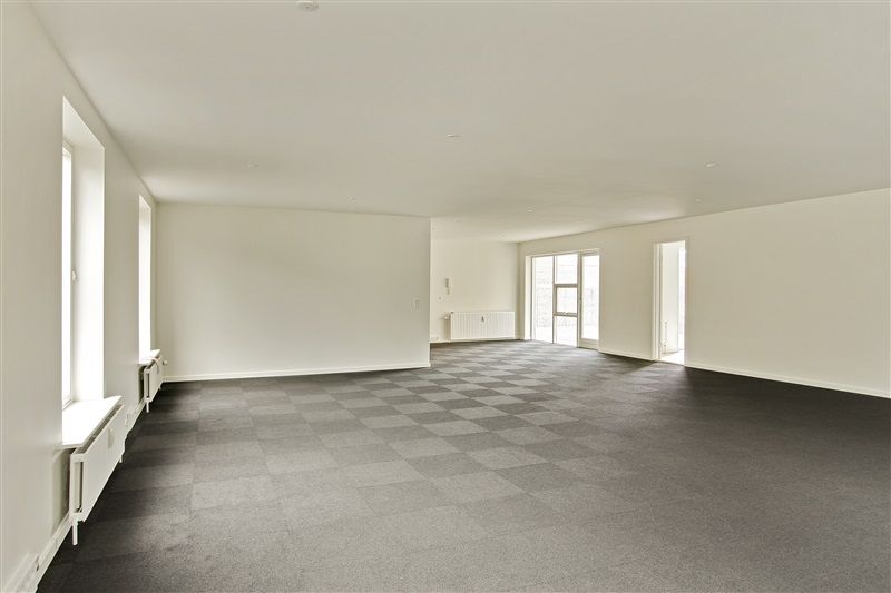 Et stort, tomt rum med krydsfinerbelagt gulv og hvide vægge og vinduer og radiatorer langs væggen