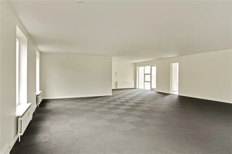 Et stort, tomt rum med krydsfinerbelagt gulv og hvide vægge og vinduer og radiatorer langs væggen