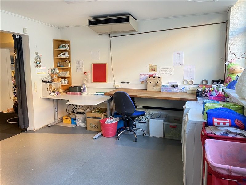 Et arbejdsværelse med to borde, stol, skraldkurve, kasser, et vasketøjsfedt, opvask og diverse materialer til håndværk