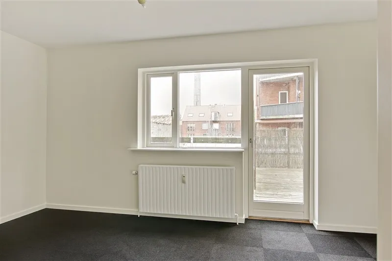 Foto af en møbleret lejlighed med lyse vægge og tæppegulv, et glasdørsystem og et hvidt radiator