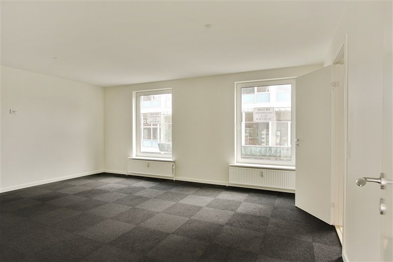 Store, tomme rum med hvide vægge og gulve dækket af grønt og sort mønstret tæppe. To vinduer med hvide rammer og skilte og