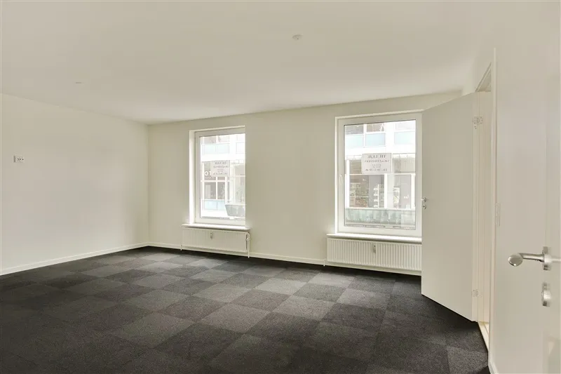 Store, tomme rum med hvide vægge og gulve dækket af grønt og sort mønstret tæppe. To vinduer med hvide rammer og skilte og