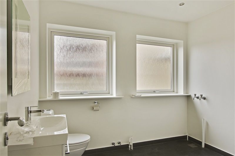 Modernt badeværelse med to vinduer, hvide vægge, sort gulv og et toilet, hvor toiletpapirhåndtaget er monteret på væggen
