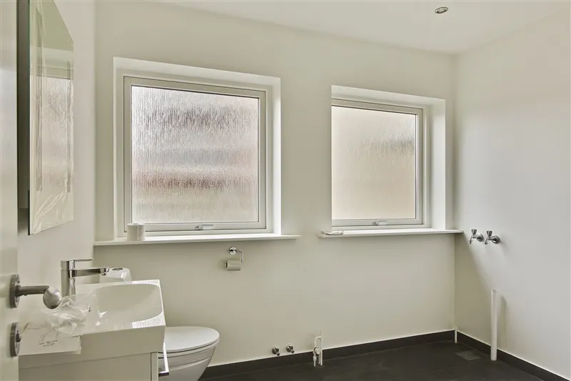 Modernt badeværelse med to vinduer, hvide vægge, sort gulv og et toilet, hvor toiletpapirhåndtaget er monteret på væggen