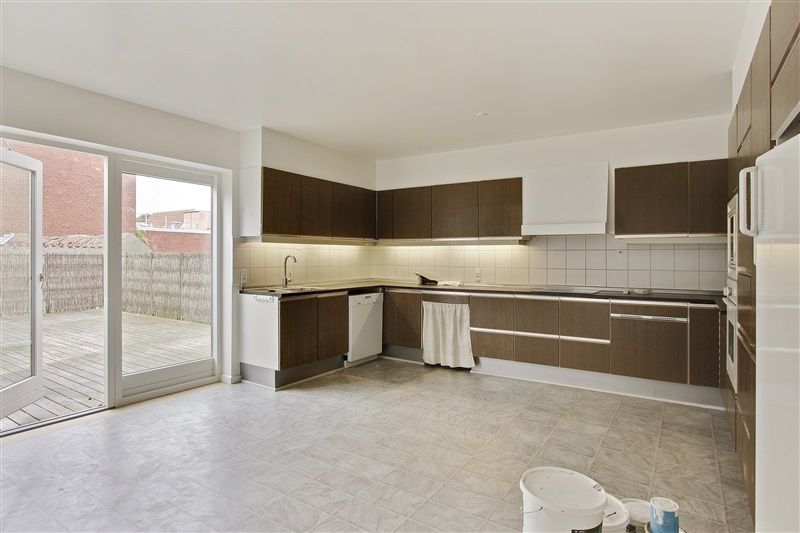 En moderne køkken med lyse og neutrale farver, hvide vægge og møbler og højloft. Døren og vinduet er åbne og giver udsigt