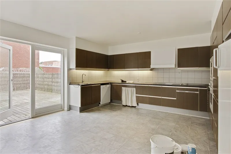 En moderne køkken med lyse og neutrale farver, hvide vægge og møbler og højloft. Døren og vinduet er åbne og giver udsigt