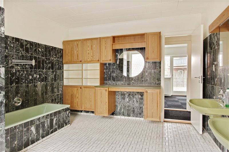 Et moderne badeværelse med marmorfliser og grønne træskabe og vandhaner. To lyseblå vasker og en stor spejl er også synlige.