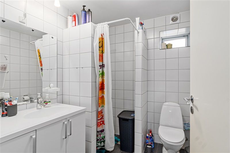 Hvidt badeværelse med toilet, brusekabine, håndvask og rengøringsprodukter samt vinduesrum med udsigt