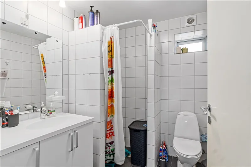 Hvidt badeværelse med toilet, brusekabine, håndvask og rengøringsprodukter samt vinduesrum med udsigt