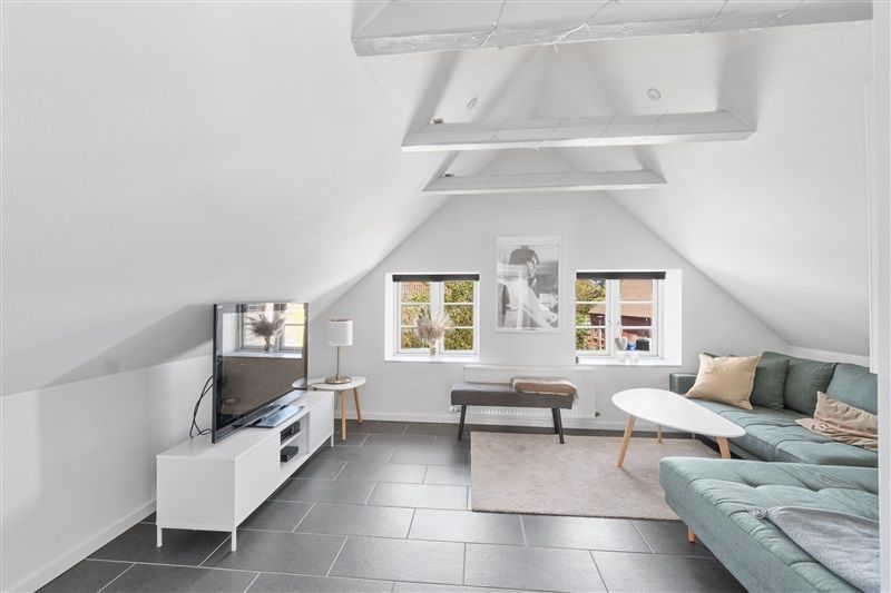 Loftsværelse med højt loft, lysegule vægge og moderne interiør med grøn sofa og tæppe. Tv, lampe og spisestuebord med vindue