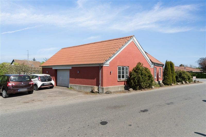 To små biler parkeret foran et rødt hus med garage og lyserøde tagfliser og skarpe hjørner