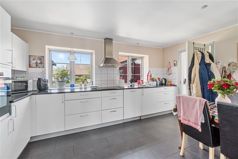 En moderne, totalrenoveret køkken med hvide skabe og lysegrå gulvfliser samt en blomstervase med røde blomster