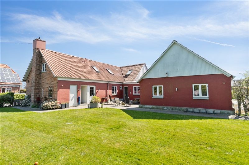 Solcellepaneler på tag, stort græsområde og rødt hus med hvide ruder og døre. Have med grønne planter og træer, hvor sofa og