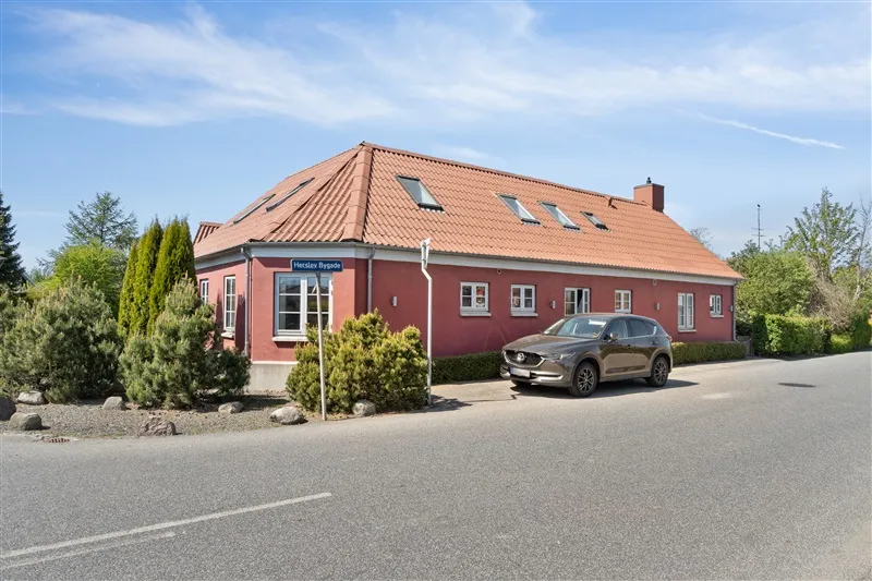 En brun og rød villa i Herslev Bygade med en parkeret bil foran og små buske og træer i forgrunden