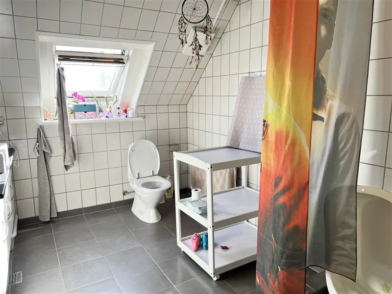 Et rent og pænt badeværelse med hvide kakeløg og en toilet med toiletpapir. Et vinduesviske og plante på vindueskarm og et