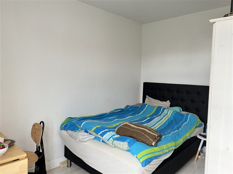 Det er et tomt soveværelse med en hvid væg og et enkelt enkeltseng med hvidt sengetøj og blå tæppe