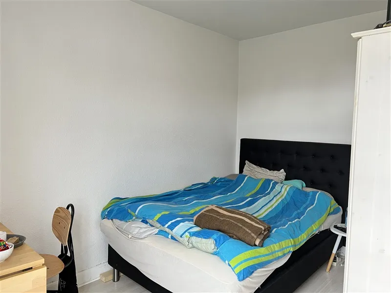 Det er et tomt soveværelse med en hvid væg og et enkelt enkeltseng med hvidt sengetøj og blå tæppe