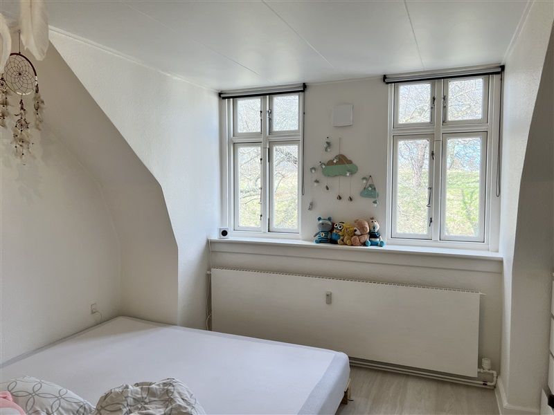 Lille børneværelse med hvide vægge, to vindue med udsigt til haven, en hvid seng med tøj og to tæpper, radiator og et